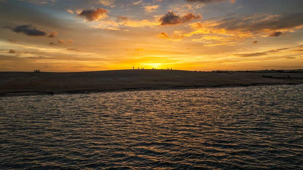 Entardecer sobre as dunas e a água, com silhuetas ao longe