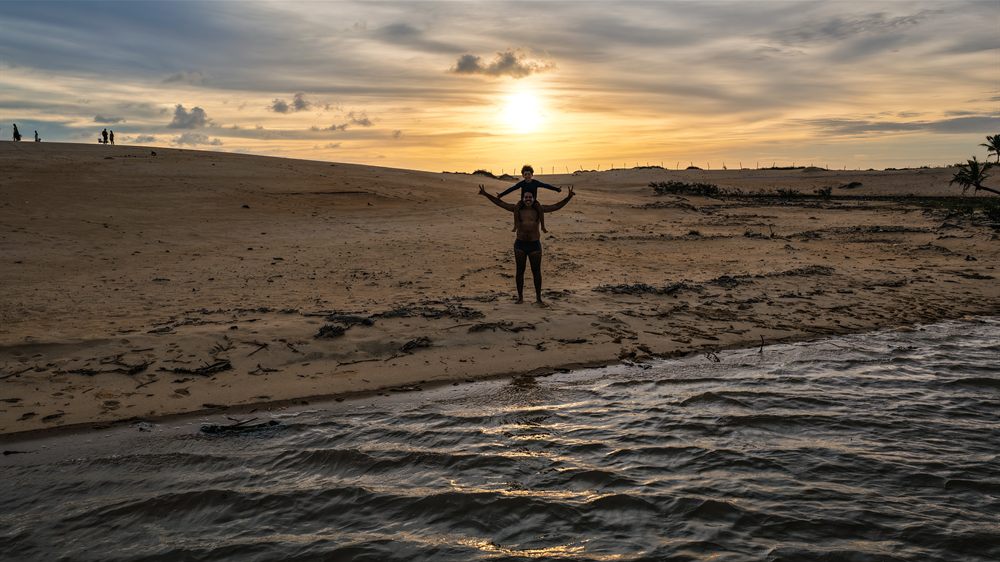 Pai com criança nos ombros nas dunas, ao pôr do sol